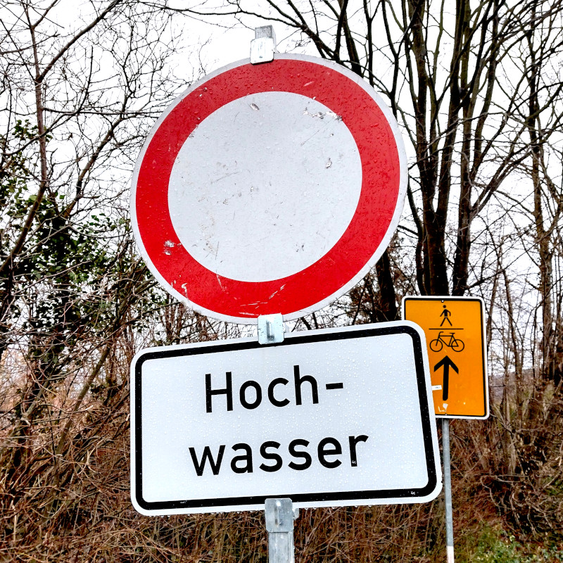 Verkehrszeichen "Verbot für Fahrzeuge aller Art" mit dem Zusatz "Hochwasser". Im Hintergrund einige Bäume und Sträucher.
