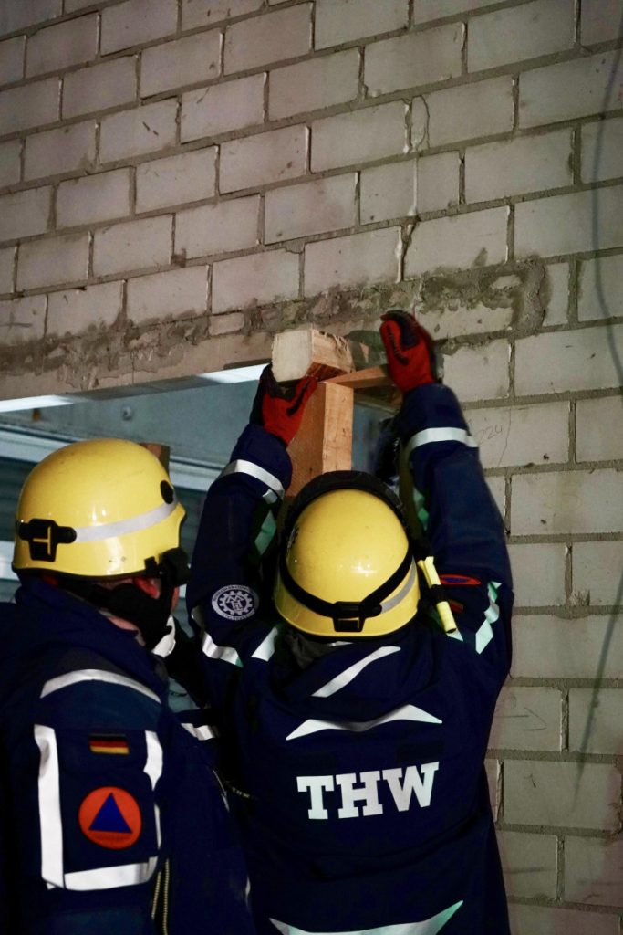 Zwei Personen in Einsatzbekleidung des THW verkeilen zwei Kanthölzer in einer Türöffnung.