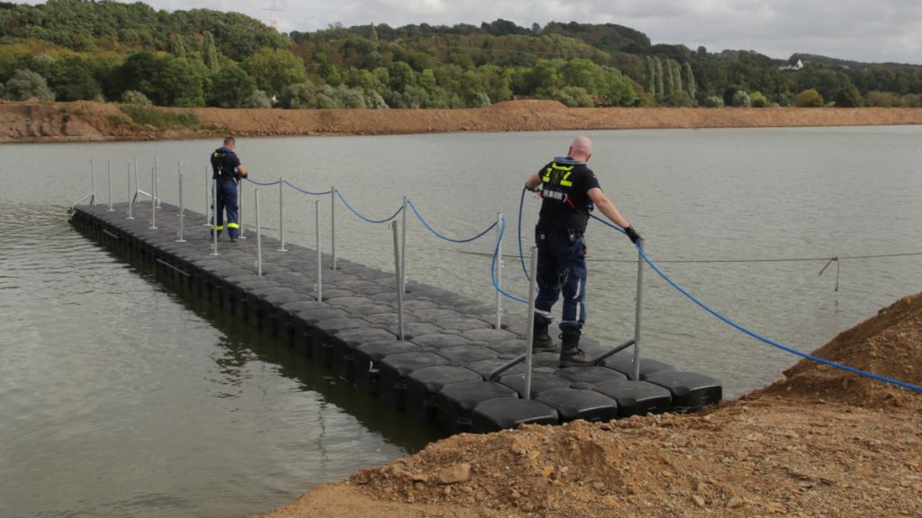Zwei Personen in Einsatzbekleidung des THW stehen auf einem im Wasser schwimmenden Steg und befestigen eine Leine als Handlauf.