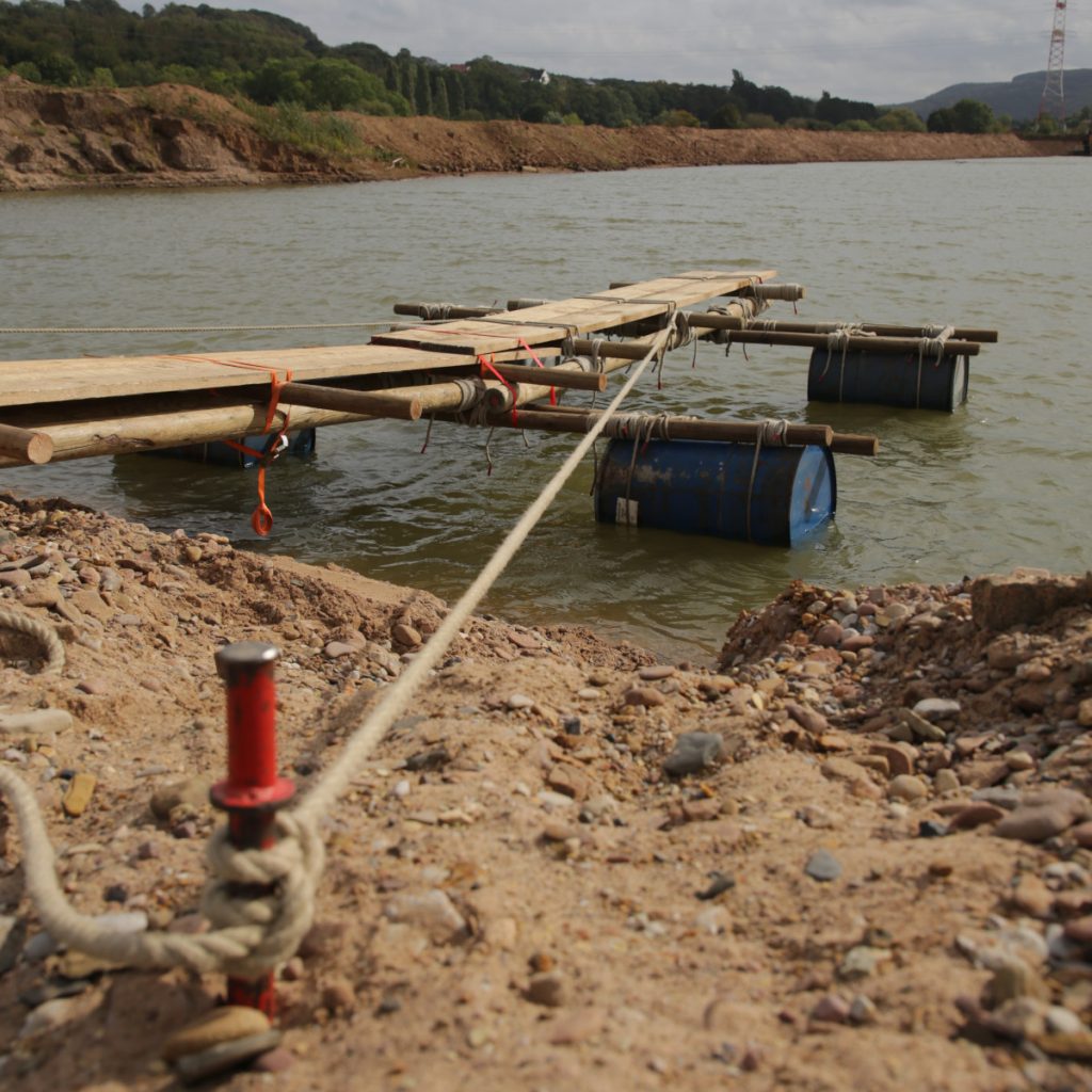 Ein Steg aus mit Leinen und Spanngurten verbundenen Hölzern, der von mehreren Metallfässern im Wasser getragen wird. Der Steg ist mit einer Leine befestigt, die in den Bildvordergrund verläuft.